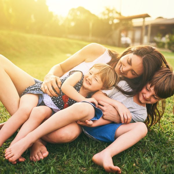 Mother, children and hug playing on grass for fun bonding in the sun outside their house in nature. Happy mom hugging kids on garden floor outdoors in playful, joy and happiness of family together.