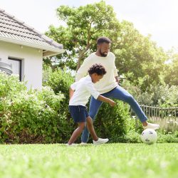 Soccer, father and happy kid on garden with sun, sports learning and goal kick together. Lawn, fun game and black family with soccer on grass with youth, sports development and bonding on field.